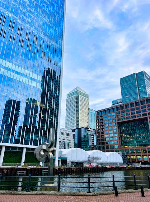 A wide view of a city skyline featuring numerous modern high-rise office buildings and skyscrapers under a blue sky with some scattered clouds. In the foreground, there is a lush green park with dense