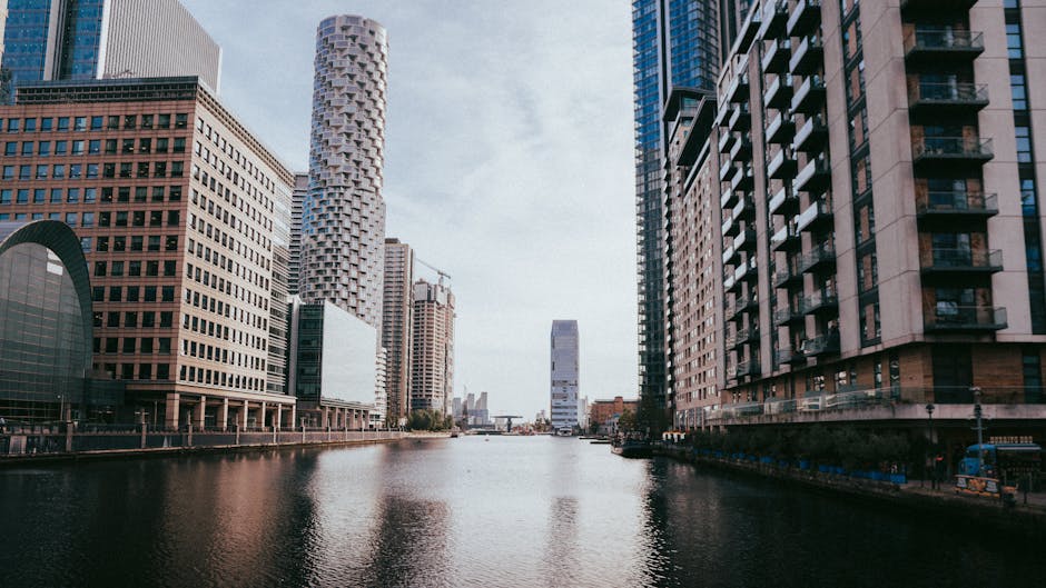 A wide-angle view of Canary Wharf in London, featuring modern high-rise office buildings along a canal. The buildings include glass towers with reflective windows and contemporary architectural designs. The canal in the foreground reflects the structures and the partly cloudy sky above. On the paved walkway along the water, there are no visible people or vehicles, but the scene is set during daylight with natural lighting. The image represents an urban environment associated with professional removals and house relocation services, illustrating the type of area where Canary Wharf Man and Van provides furniture transport and moving logistics. The scene emphasizes the setting for city-based property moves and furniture loading processes in a professional moving context.