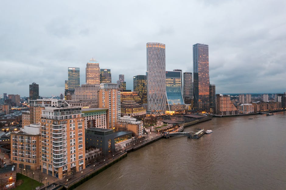 A panoramic view of Canary Wharf's skyline during evening hours, showing a cluster of tall office buildings and residential apartments with illuminated windows. In the foreground, there is a river with a boat docked along the quay, and a promenade running parallel to the water. The paved area near the water features a few cars and pedestrians, with some compound structures and outdoor lighting visible. The scene is captured with natural daylight fading into dusk, highlighting the modern architecture and urban environment. Interior shots of a property with furniture, cardboard boxes, and packing materials such as plastic wrap and blankets may be seen during a typical home relocation or furniture transport process, often involving a loading area close to the building entrance. A van from Canary Wharf Man and Van is sometimes visible nearby to facilitate the packing and moving activities, including lifting, carrying, and loading household items for a house removal service, in alignment with the Heron Quays moving checklist for Canary Wharf removals.