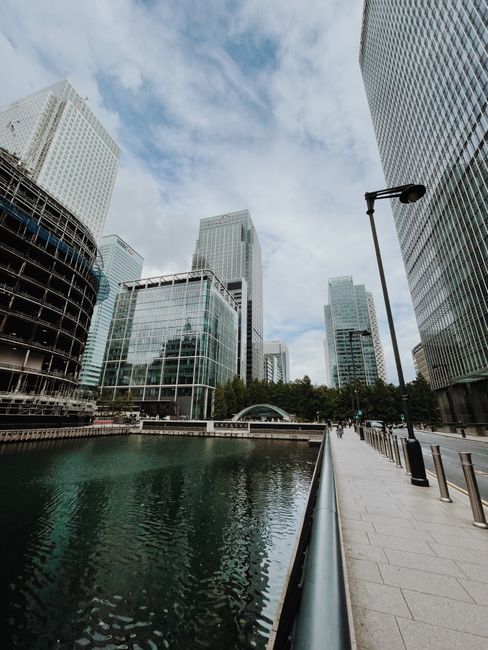 A wide-angle view of Canary Wharf in London, featuring modern high-rise office buildings along a canal. The buildings include glass towers with reflective windows and contemporary architectural designs. The canal in the foreground reflects the structures and the partly cloudy sky above. On the paved walkway along the water, there are no visible people or vehicles, but the scene is set during daylight with natural lighting. The image represents an urban environment associated with professional removals and house relocation services, illustrating the type of area where Canary Wharf Man and Van provides furniture transport and moving logistics. The scene emphasizes the setting for city-based property moves and furniture loading processes in a professional moving context.