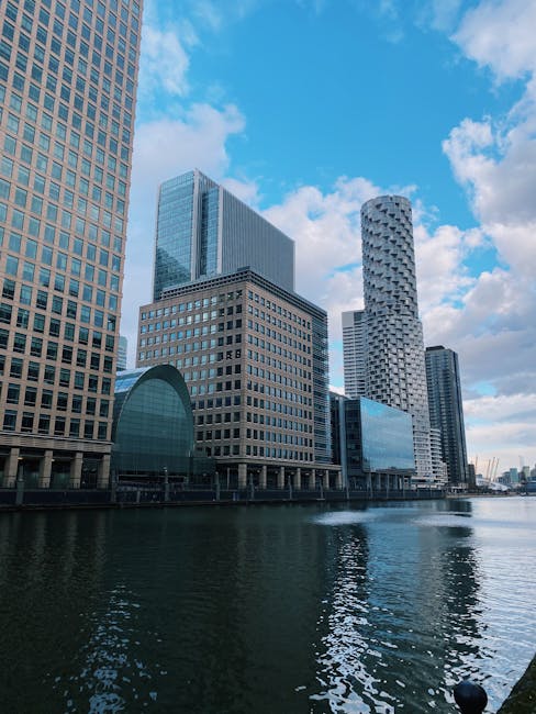 A view of modern skyscrapers along the edge of the water in Canary Wharf, with tall office buildings featuring glass facades and geometric designs. The buildings include a mixture of rectangular, curved, and cylindrical shapes, creating a striking city skyline against a partly cloudy sky. The water in the foreground reflects the buildings and sky, highlighting the urban landscape. This scene exemplifies the contemporary architecture typical of a central London business and residential district, often visited during house removals or relocation projects, with the area accessible for professional moving services such as Canary Wharf Man and Van involved in logistics and furniture transport within the estate.