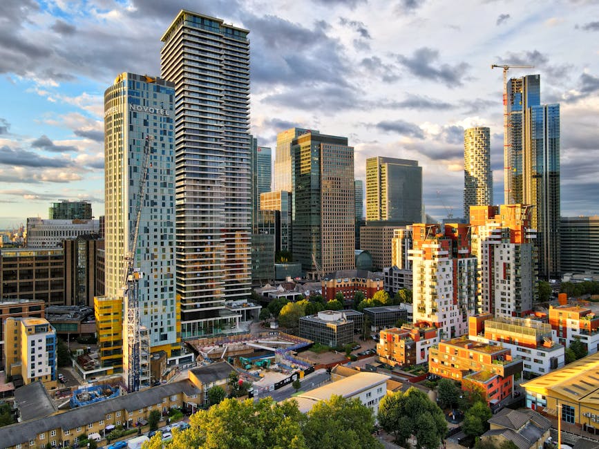 A cityscape image showing a collection of modern high-rise buildings and skyscrapers in Canary Wharf, with a mix of glass and steel facades reflecting the sky and surrounding structures. In the foreground, there are lower-rise buildings and trees, along with a construction crane beside a building under development, indicating ongoing building work. The scene is captured during late afternoon or early evening, with sunlight casting warm tones on the buildings, highlighting the details of the architecture. The area appears busy with various construction materials and equipment visible on the ground level, consistent with ongoing home relocation or furniture transport activities as part of the moving process. Canary Wharf Man and Van may be involved in managing logistics for these buildings, supporting packing, loading, and transportation services in this urban environment.