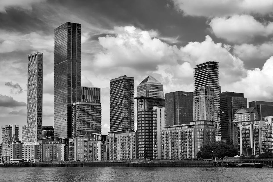 A view of modern skyscrapers along the edge of the water in Canary Wharf, with tall office buildings featuring glass facades and geometric designs. The buildings include a mixture of rectangular, curved, and cylindrical shapes, creating a striking city skyline against a partly cloudy sky. The water in the foreground reflects the buildings and sky, highlighting the urban landscape. This scene exemplifies the contemporary architecture typical of a central London business and residential district, often visited during house removals or relocation projects, with the area accessible for professional moving services such as Canary Wharf Man and Van involved in logistics and furniture transport within the estate.
